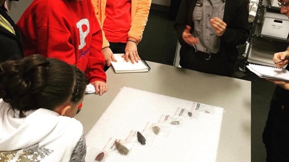 A group of excited students surround a table of arrowheads laid out in a sequential line.
