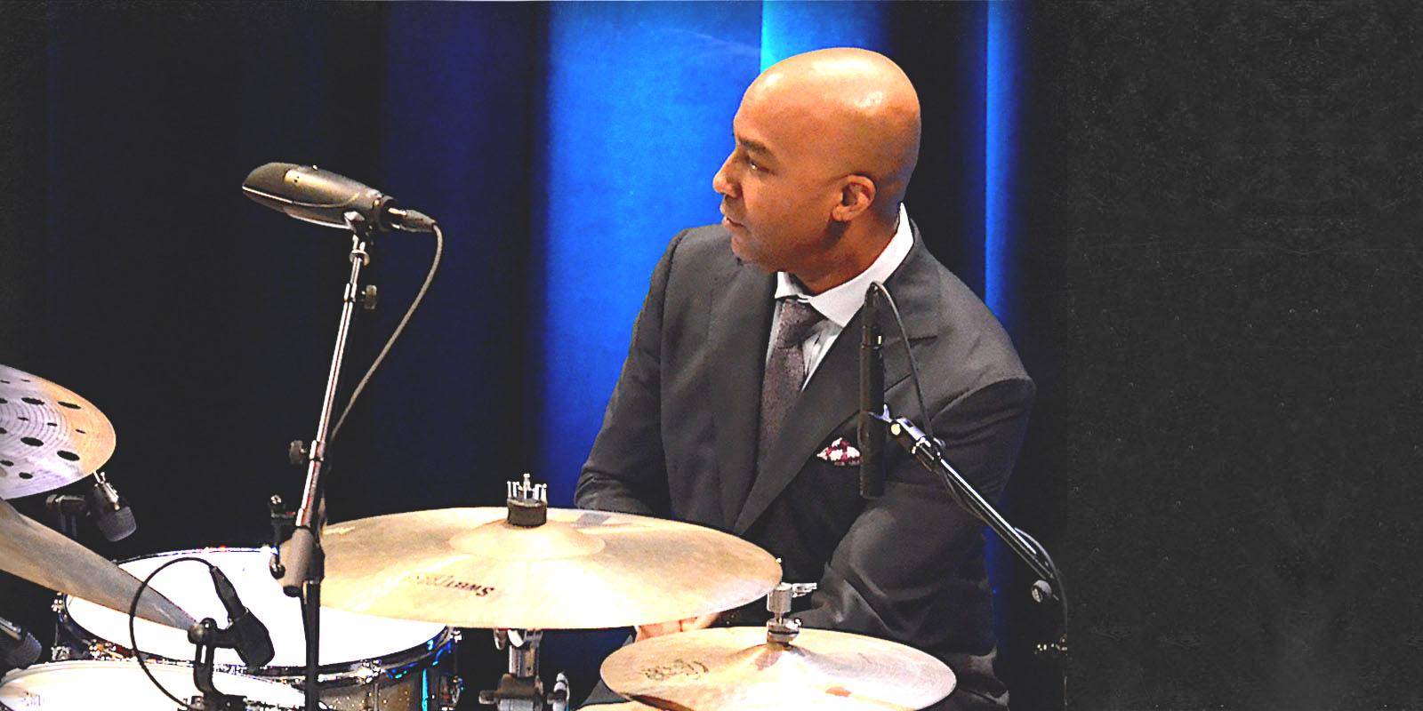A man dressed in a suit sits behind a drum set