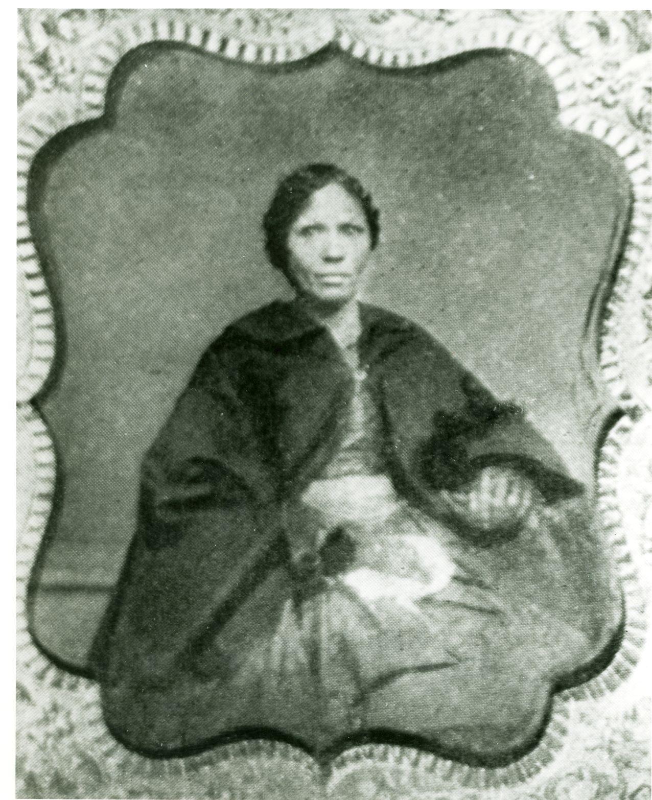 A woman sitting wearing a historic period dress and black shawl.