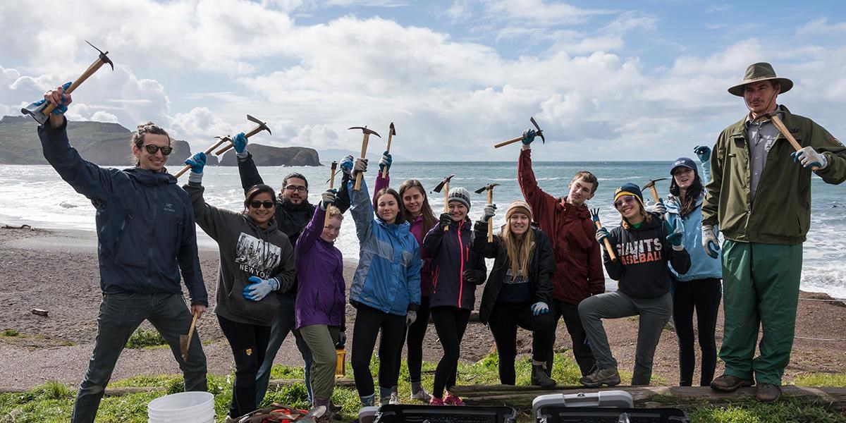 A group of volunteers celebrating holding up their tools in front of the beach