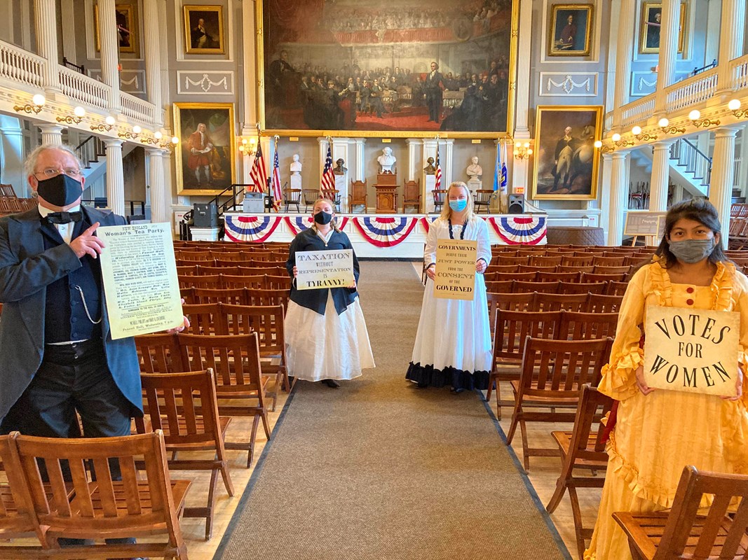 Four people dressed in 1870s period clothing standing in the great hall of Faneuil Hall.