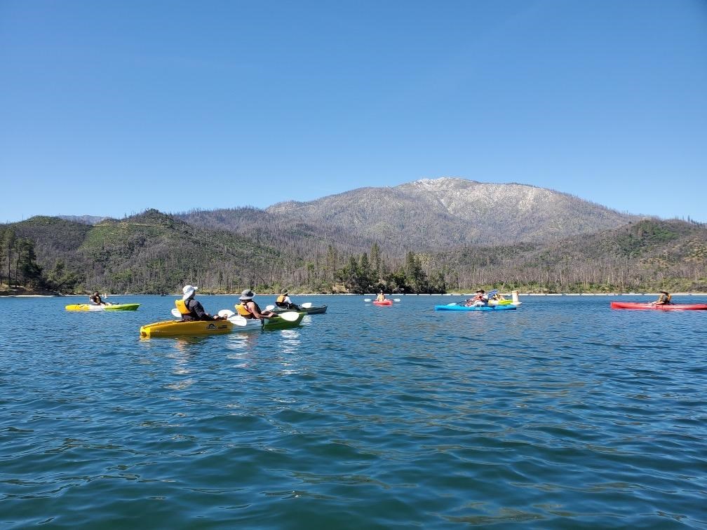 Blue sky and blue water with a tall mountain in the background. People in kayaks paddling away.