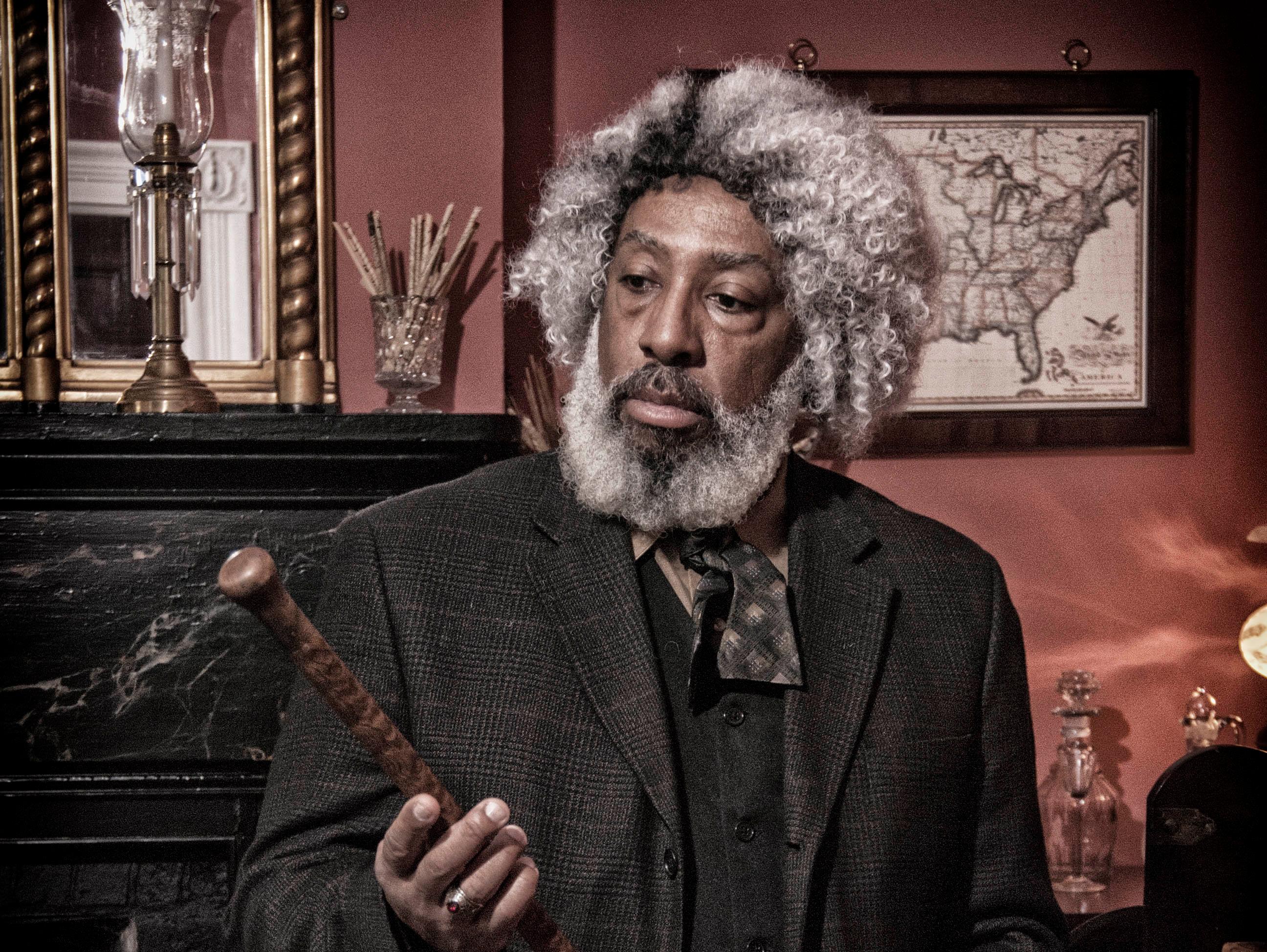 A  man with a gray beard and hair, wearing a jacket, vest, and cravat, inspects a wooden staff.