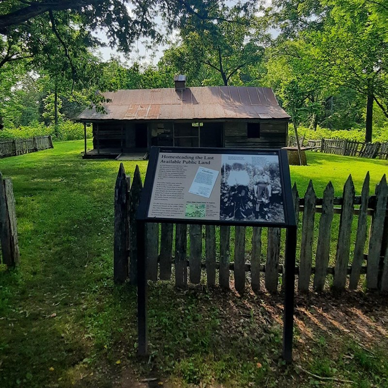 Color photograph of an informational sign in front of a wooden fence and a historic wooden house.