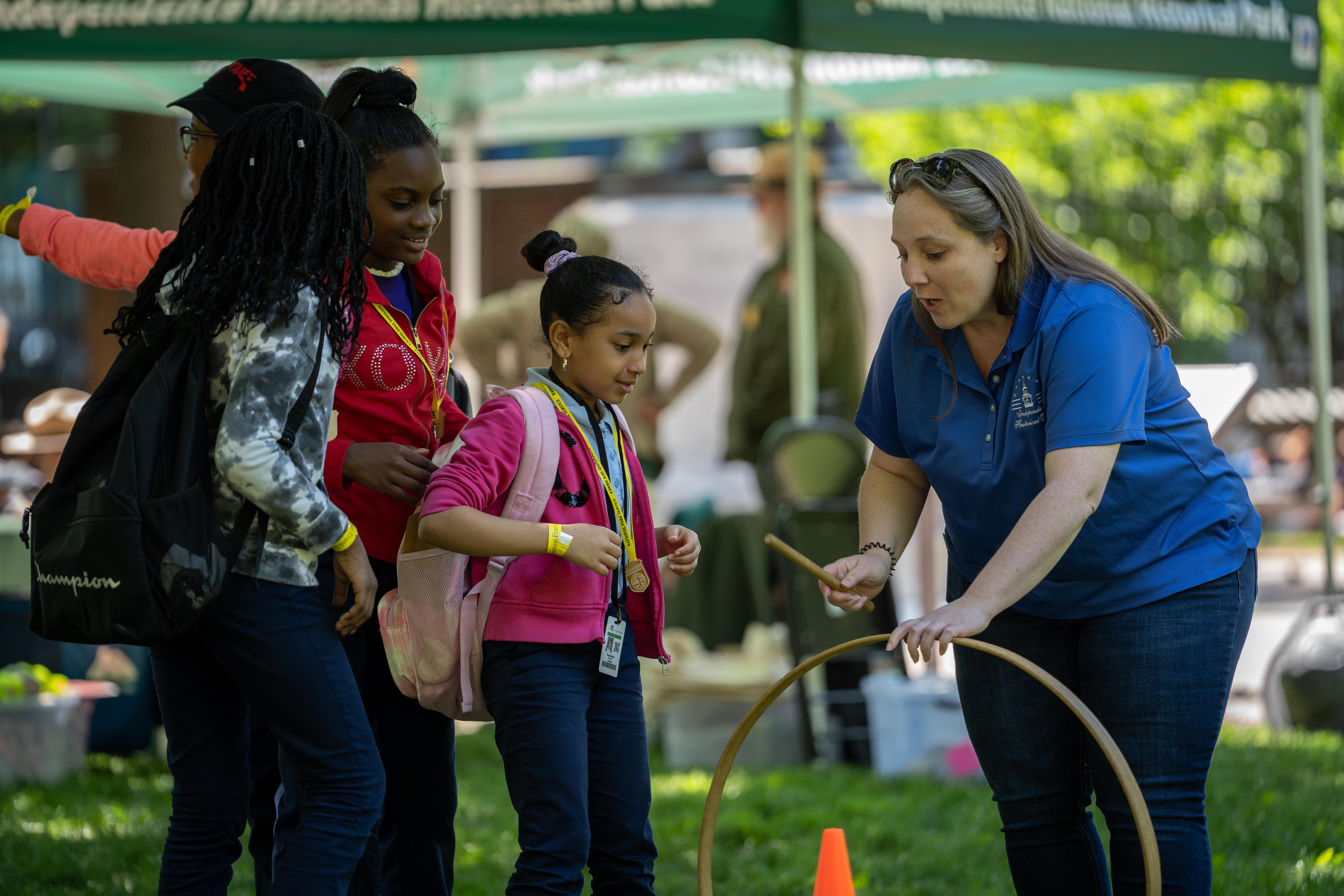 Children line up excitedly while a woman hands a girl a hoop and stick.