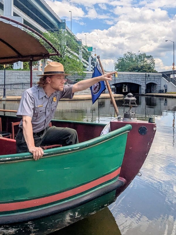 A uniformed park ranger pointing from the bow of a small, green canal boat.