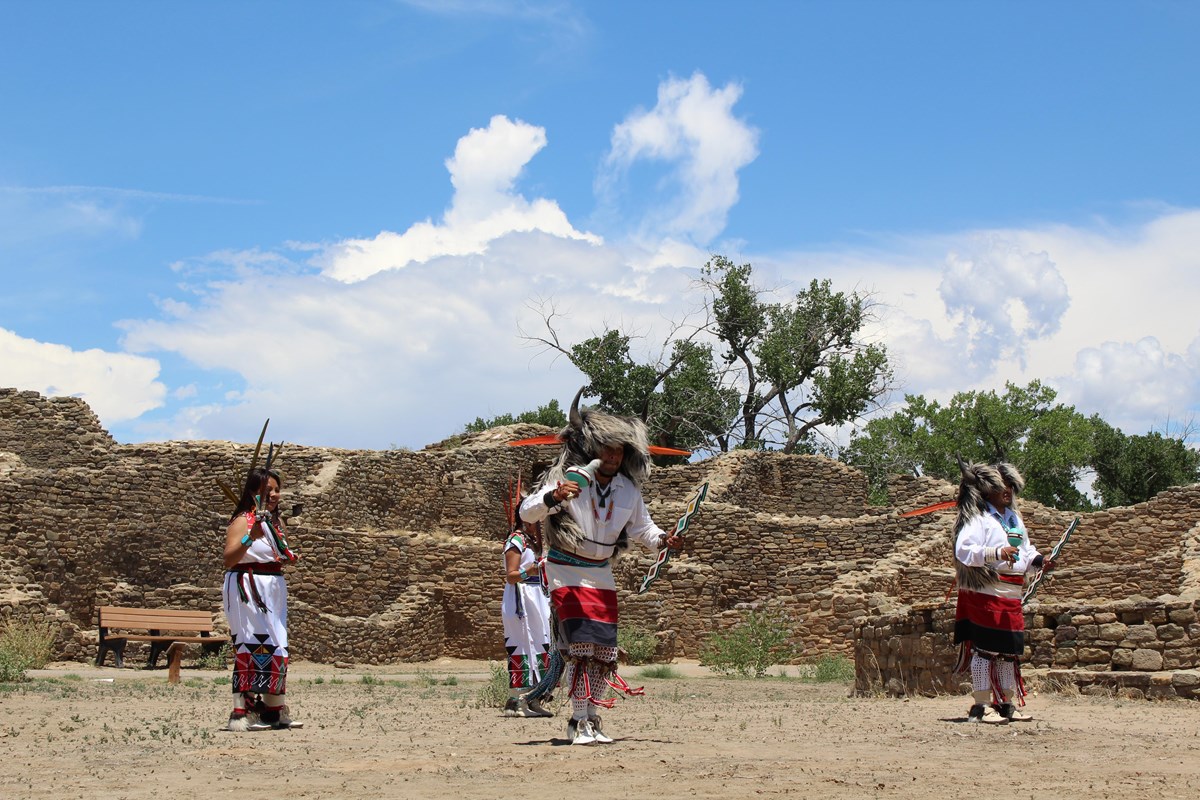 Tribal dancers perform amidst stone ruins, with green trees and arching clouds in the background.