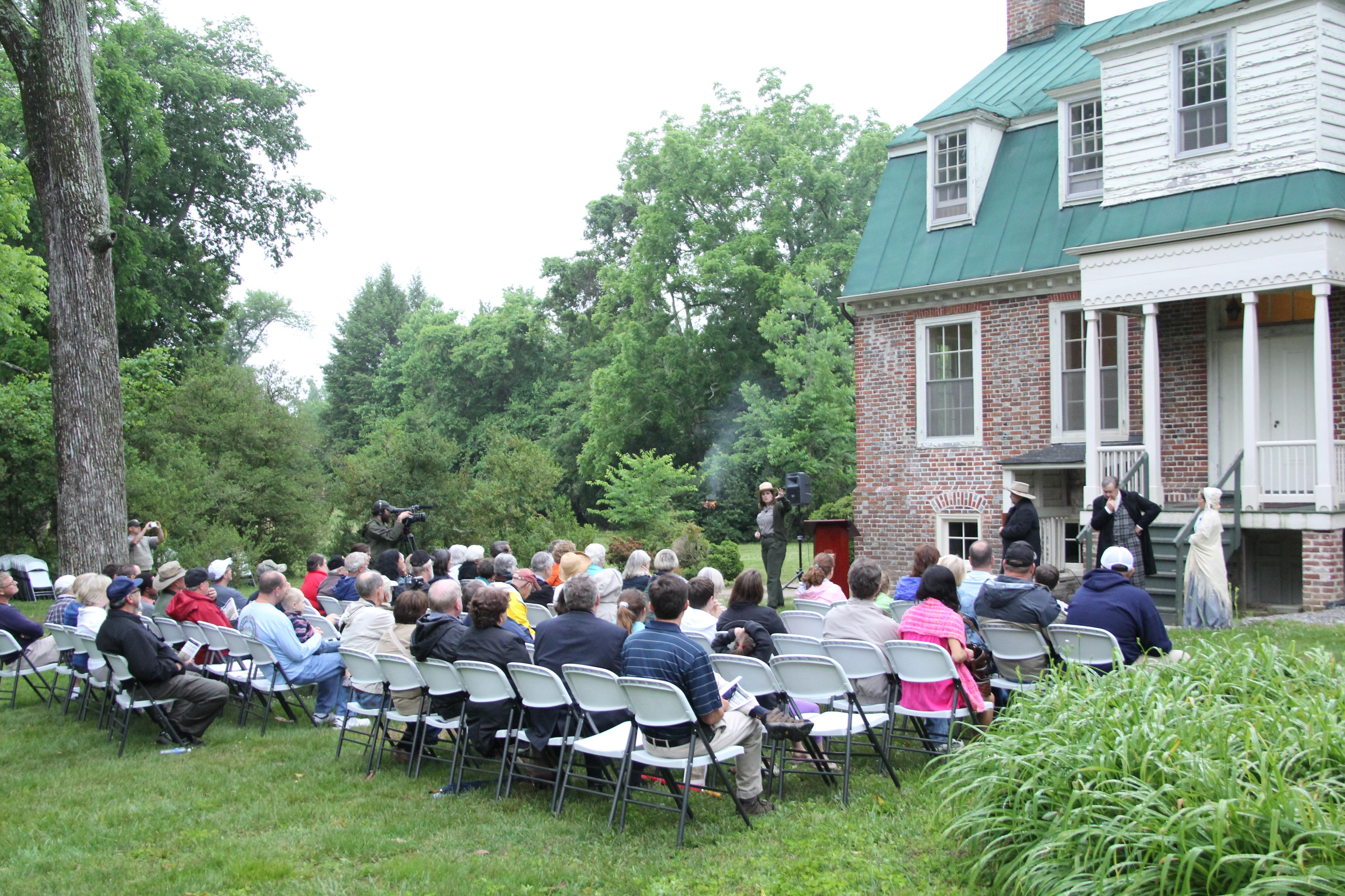 an audience seated outdoors in white folding chairs in front of an 18th-century brick mansion.