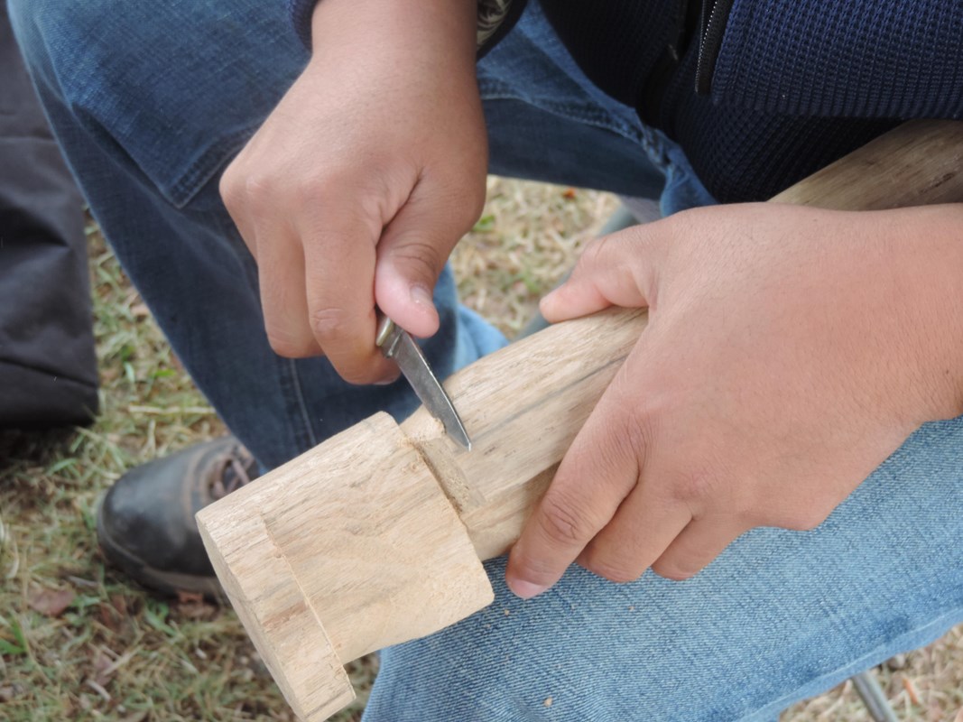 A closeup of two hands carving a piece of wood with a pocketknife.