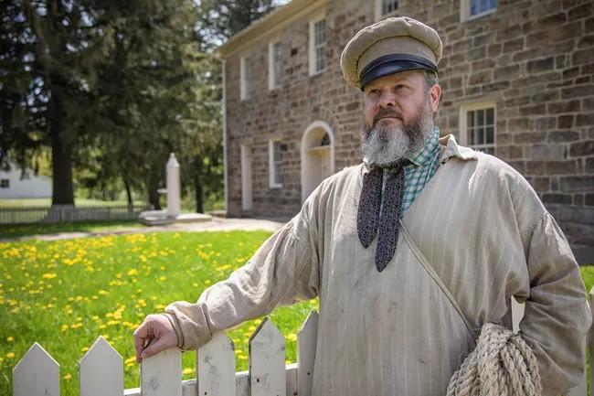 A man in period clothing holds rope in front of a stone tavern.