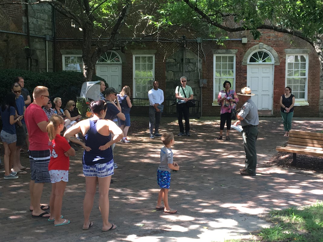 Uniformed park ranger conducting tour for semicircle of people under mottled shade on a sunny day.