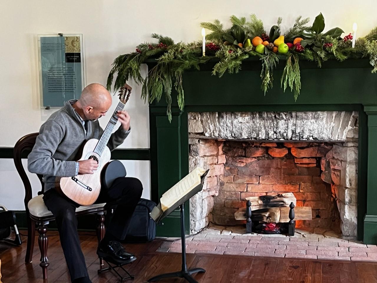man seated playing guitar next to fireplace. Mantel decorated with greenery.