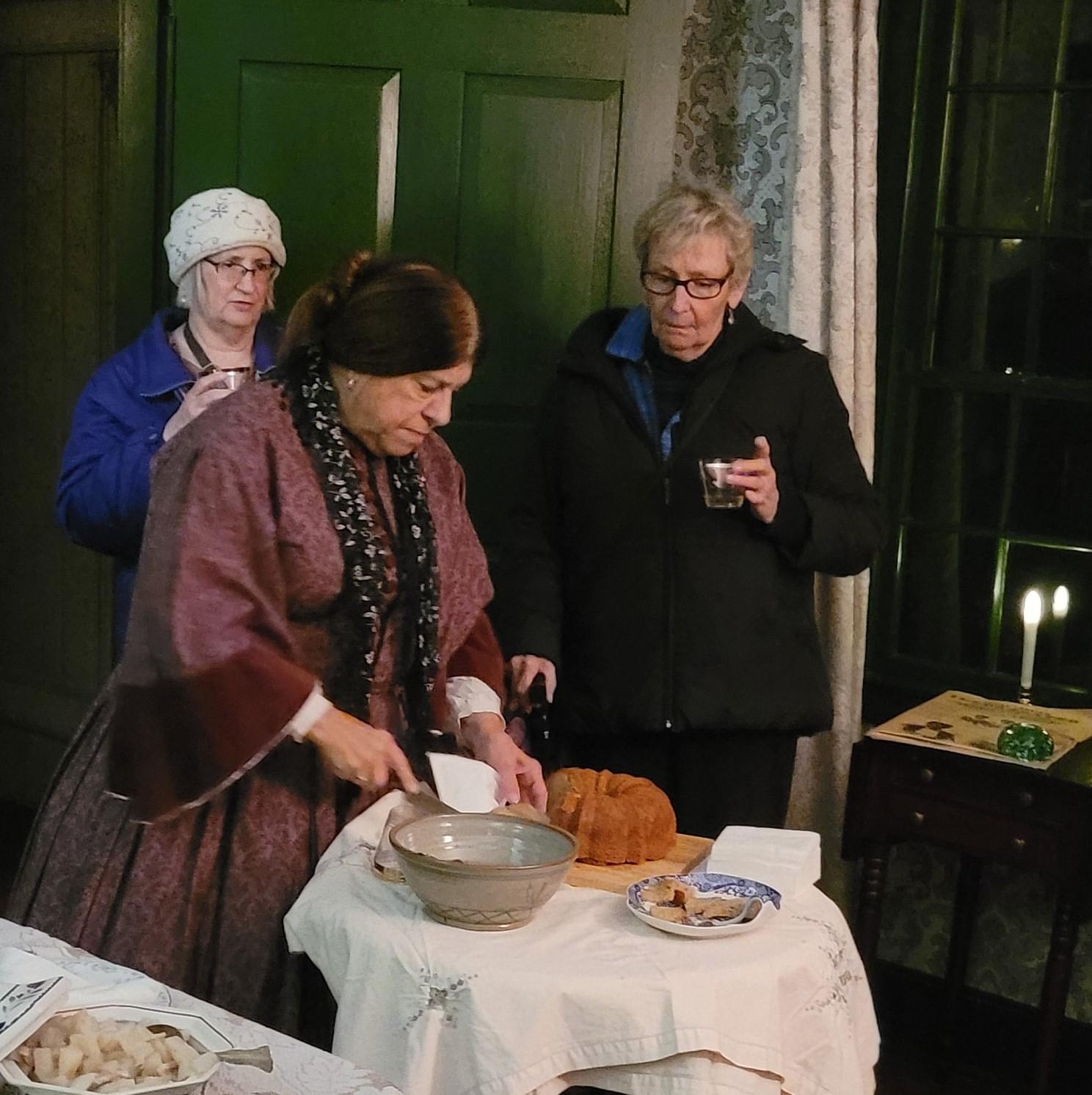 woman in 19th century dress at small table with punch bowl and cake. Two visitors behind her.