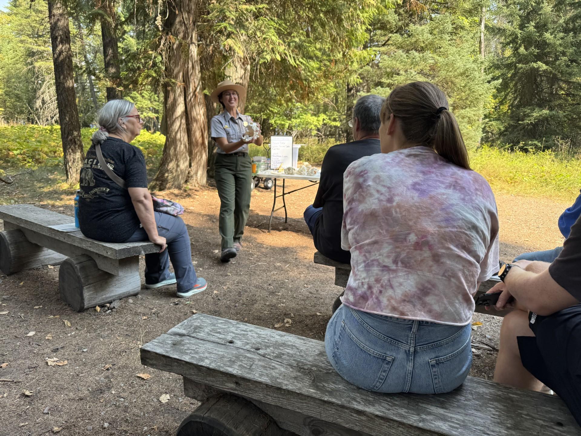 Park ranger holds up an animal skull in front of a table and trees.