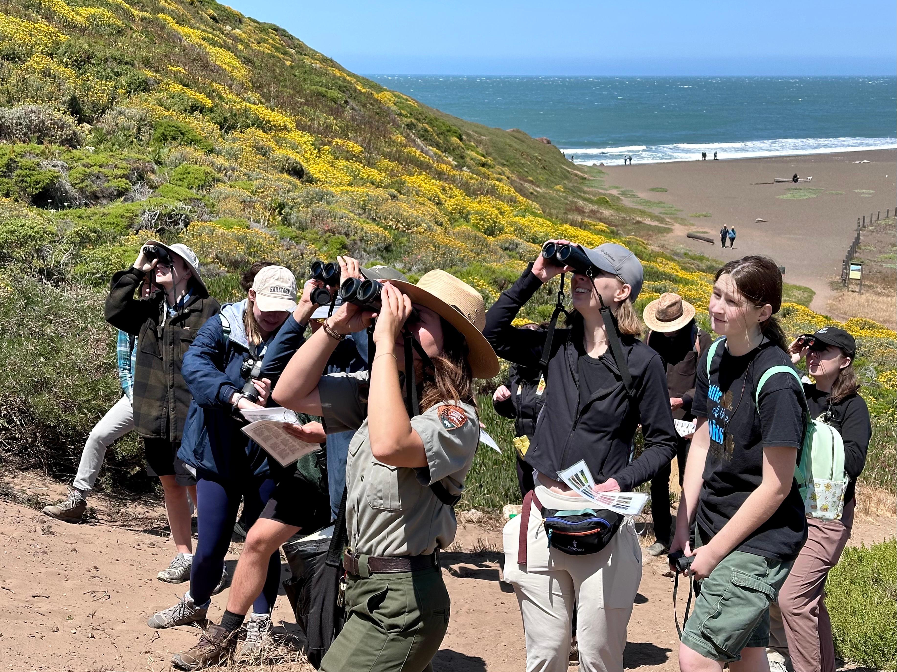 a ranger in uniform looks up with binoculars and a group of visitors around her do the same