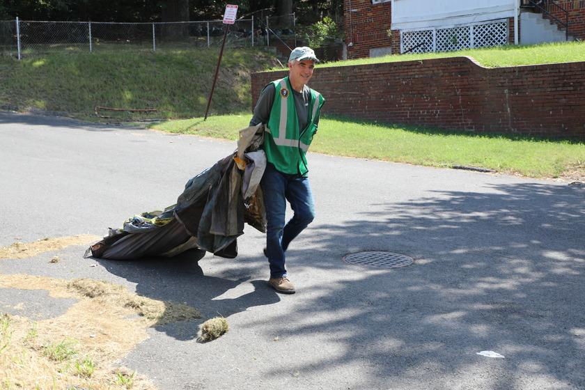 A person wearing a green safety vest dragging a discarded tent behind them near a grassy hill.