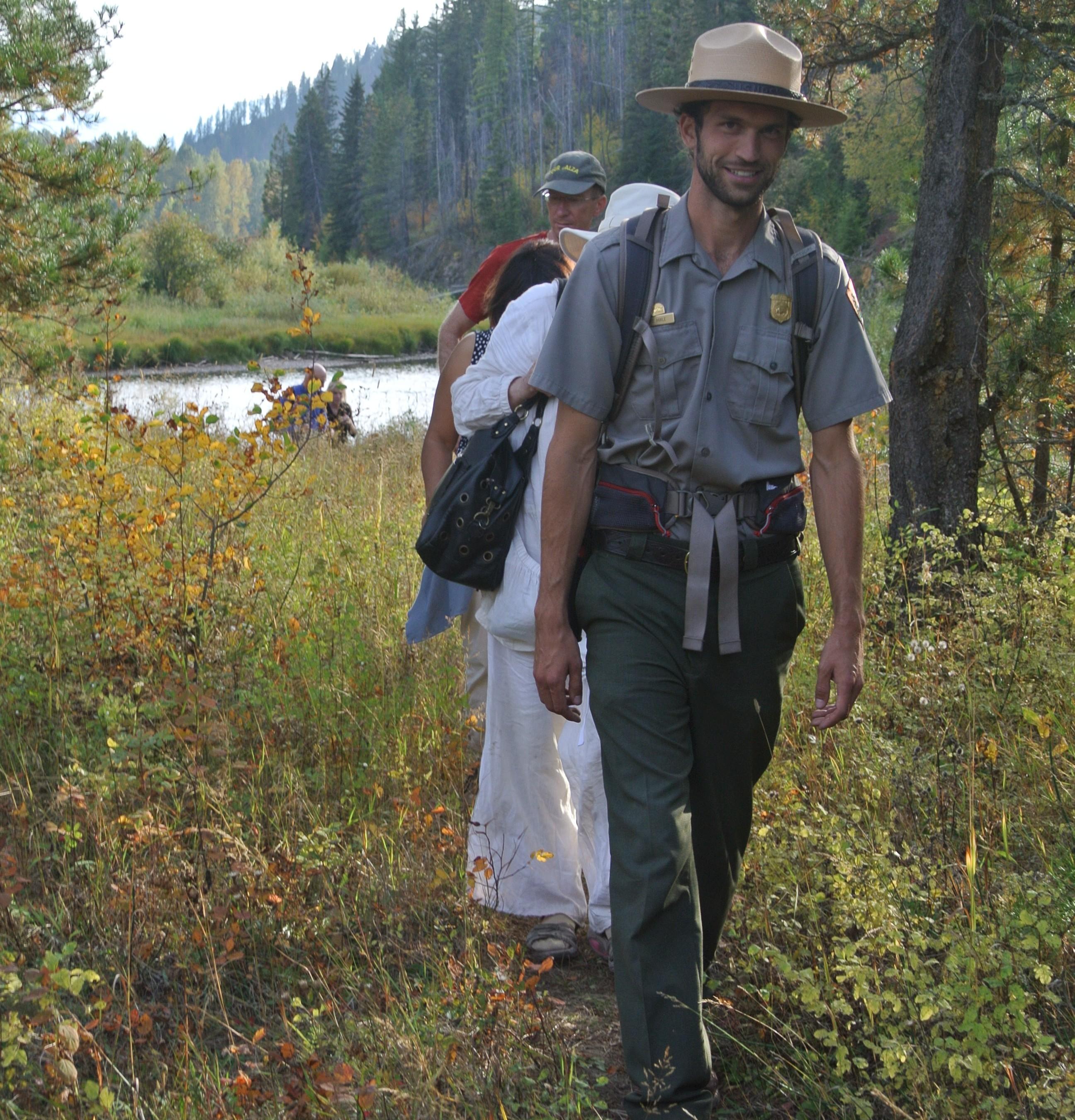 Park ranger and people walking on trail with trees and creek behind them.