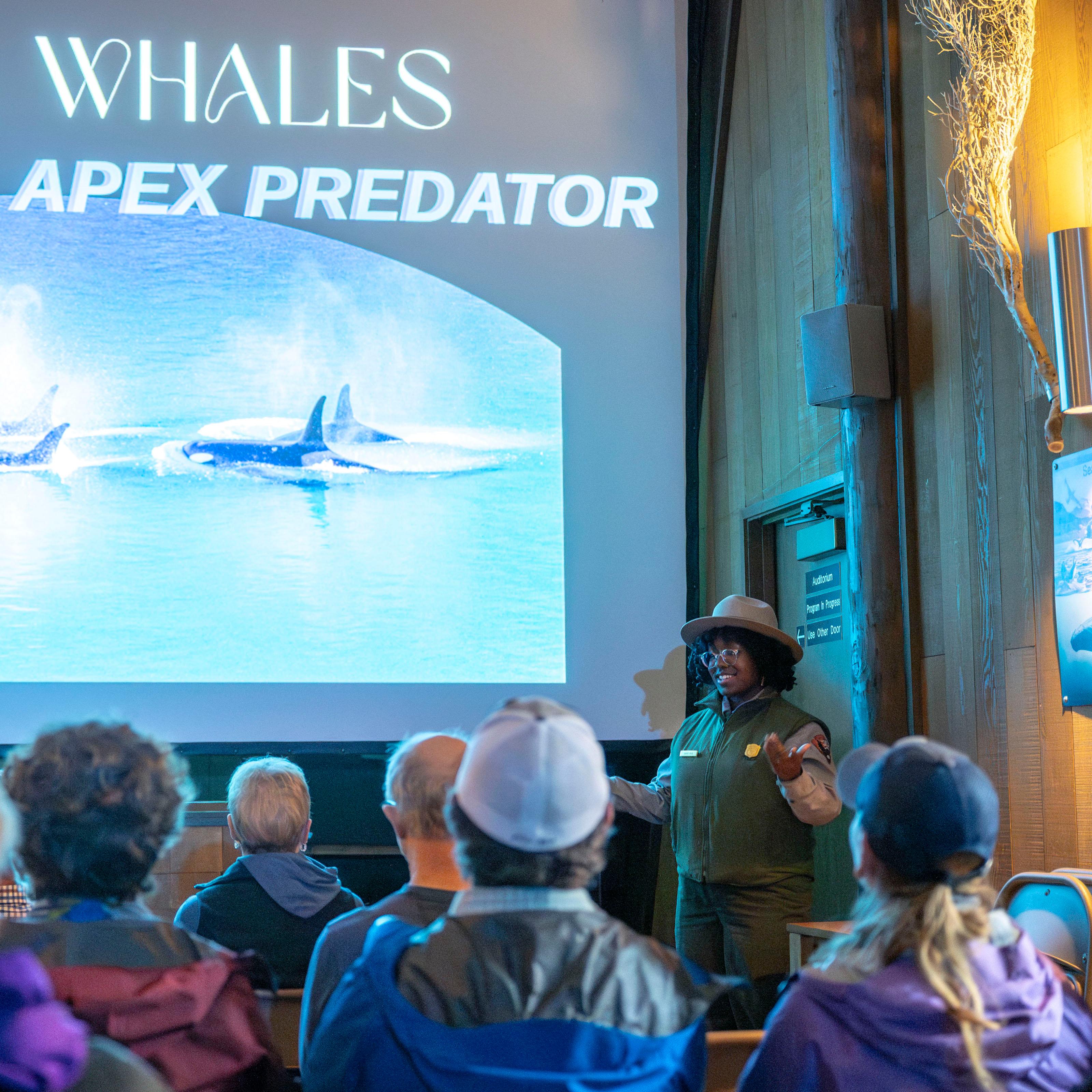 A park ranger speaks to a theater full of people while showing a graphic on a projector screen.
