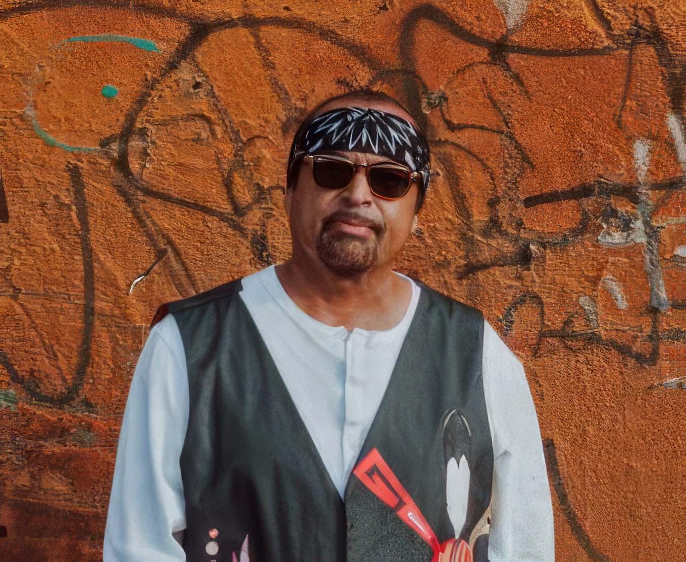 A middle-aged Native American Man wearing sunglasses stands against a brown wall.