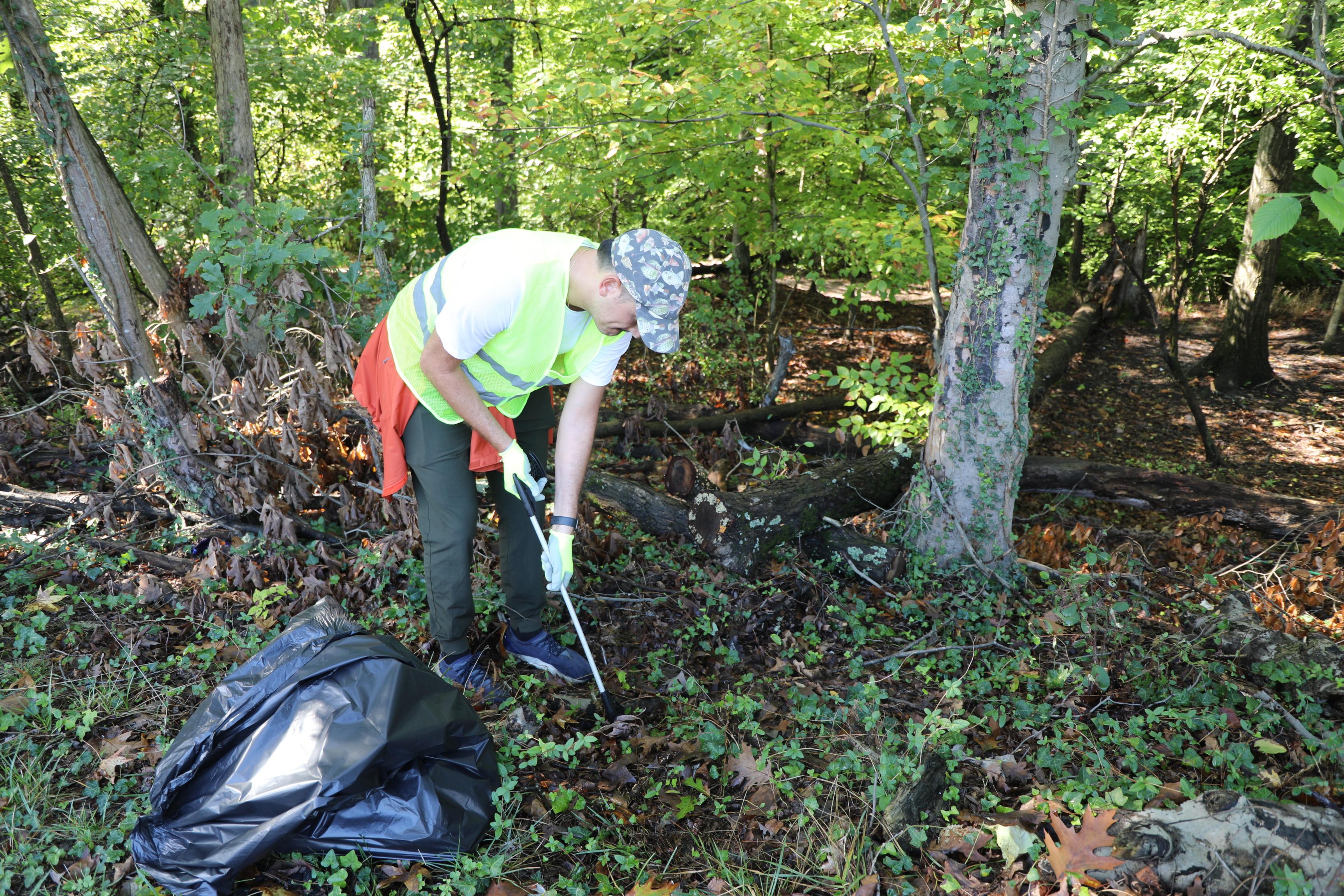 A volunteer in a neon vest uses a long grabber tool to pick up litter from a leafy forest floor.
