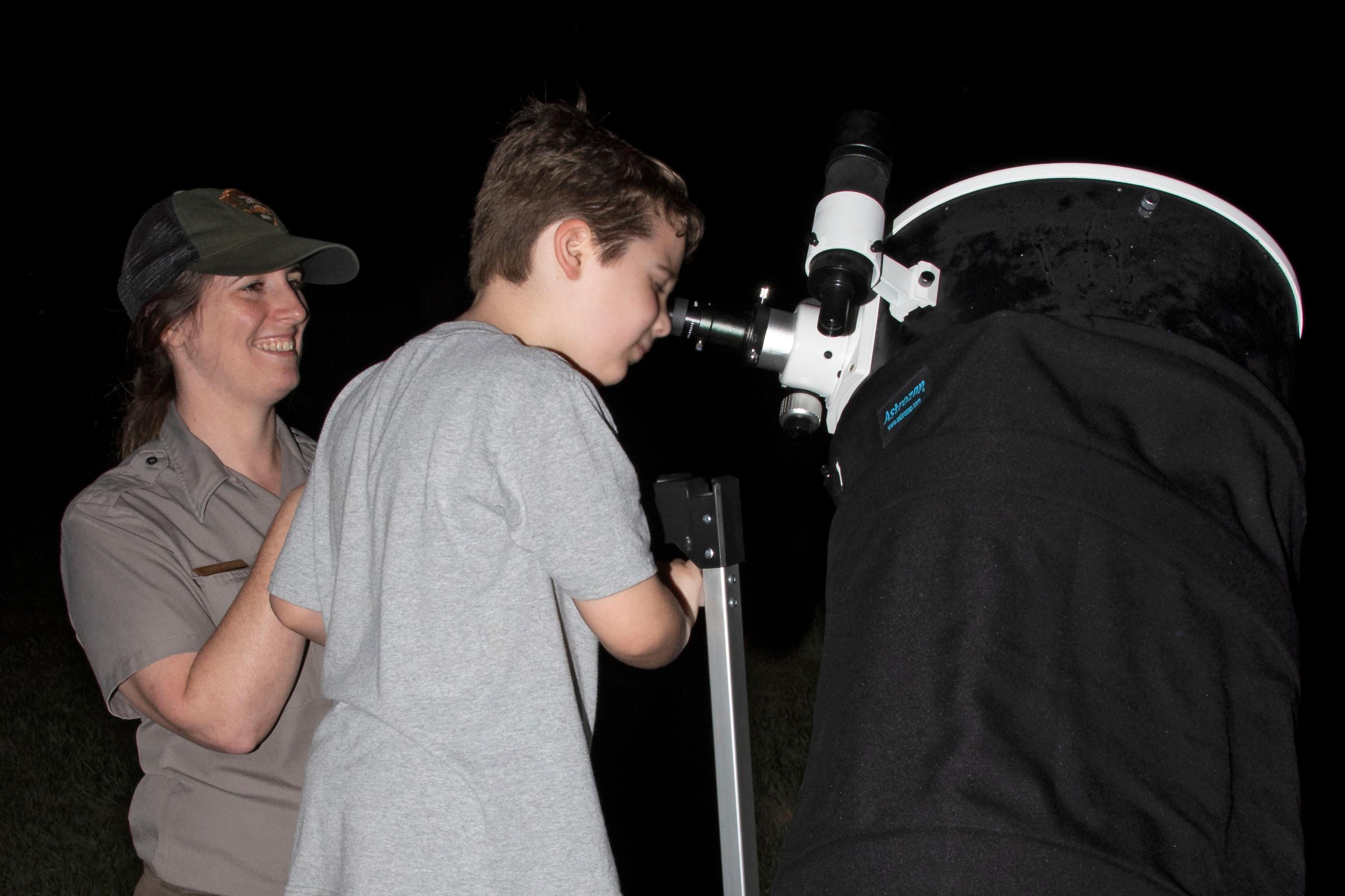 A ranger stands to the side while a young boy looks through a large telescope.