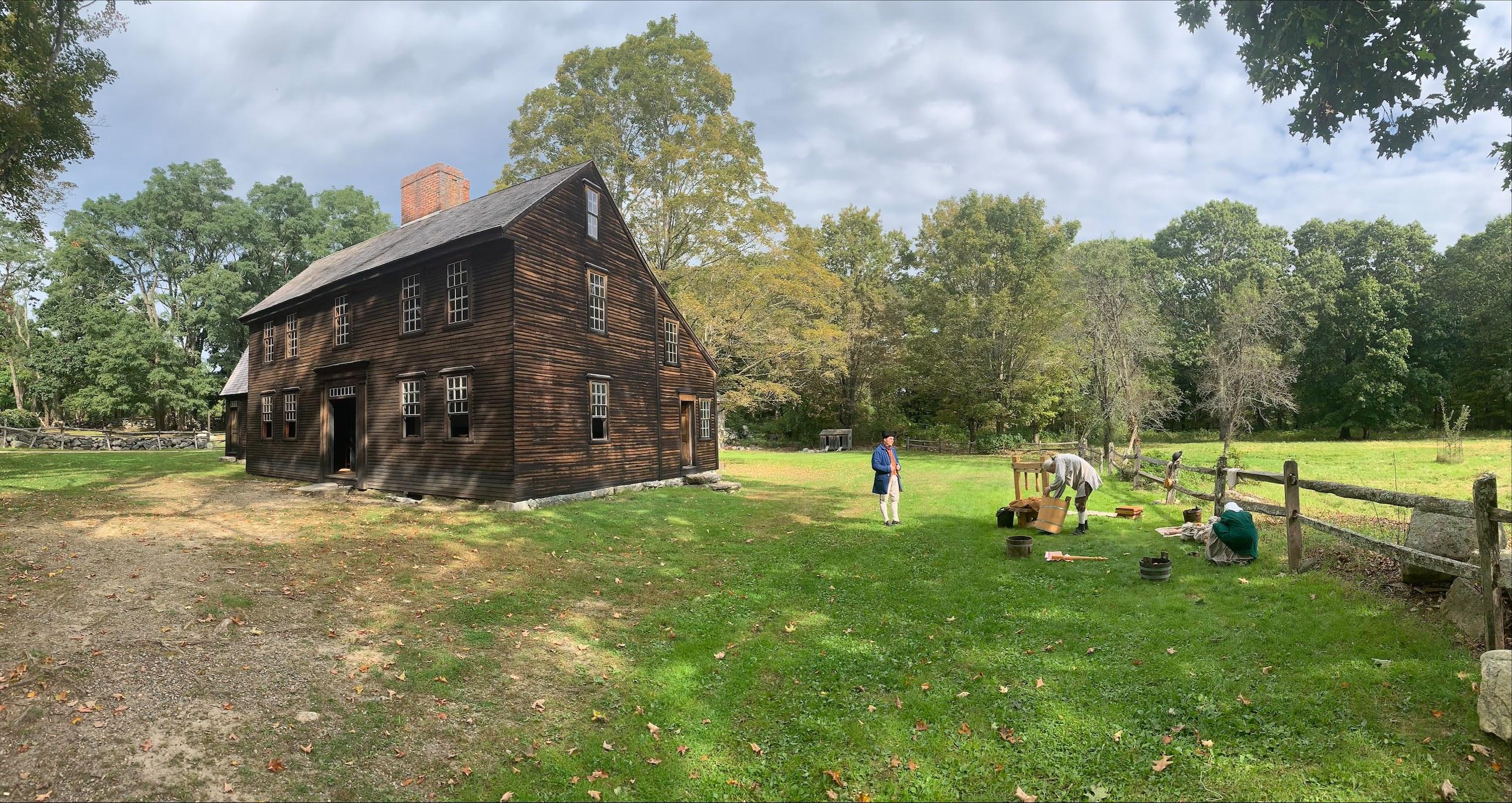 A historic wooden house in a field with people dressed in colonial clothing outside, and a cider pre