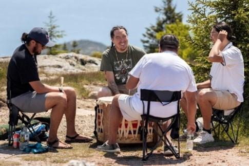 Four men seated and singing in a circle around a large drum with trees, rocks, and mountain summits