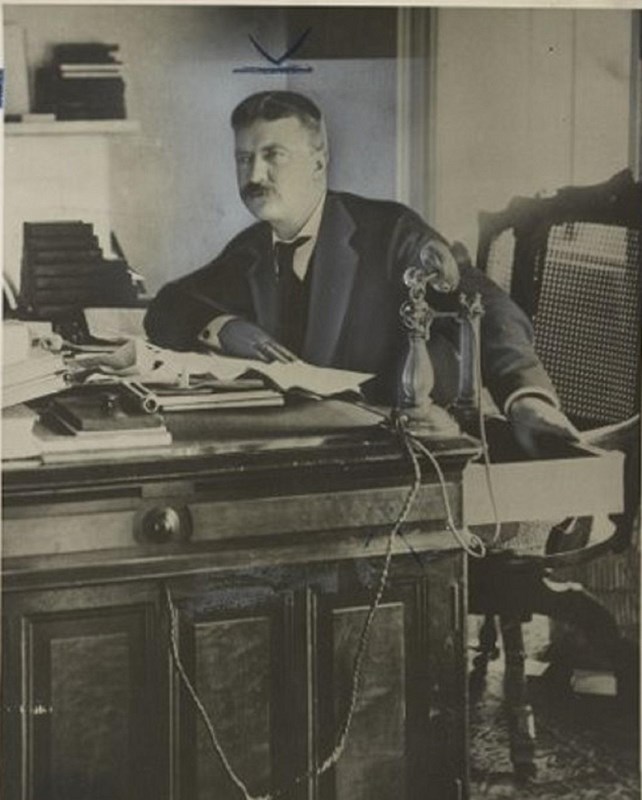sepia photograph of Theodore Roosevelt sitting at his desk,  learning forward with his right elbow