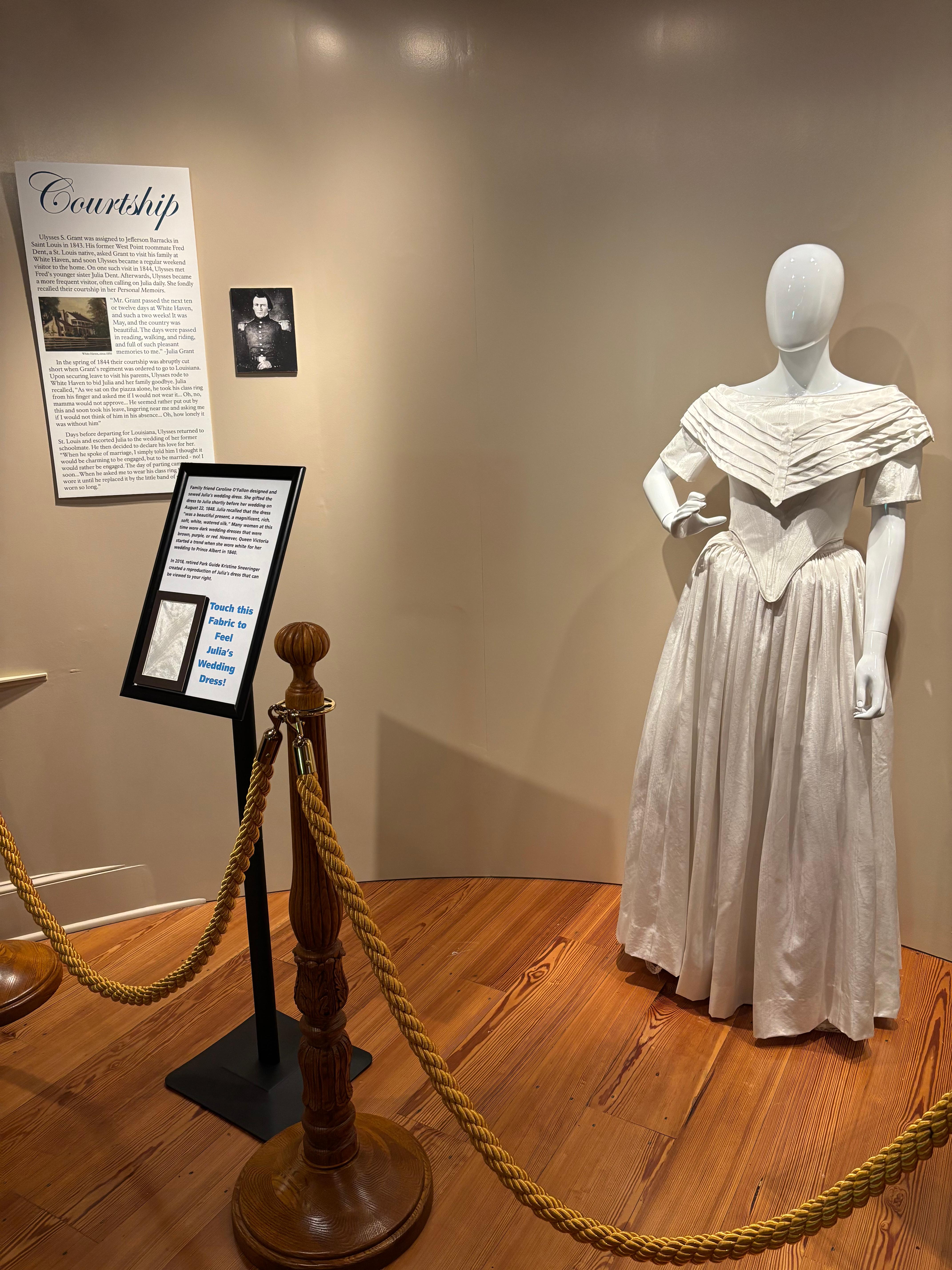 White wedding dress on mannequin in front of tan wall. Exhibit panels adjacent.