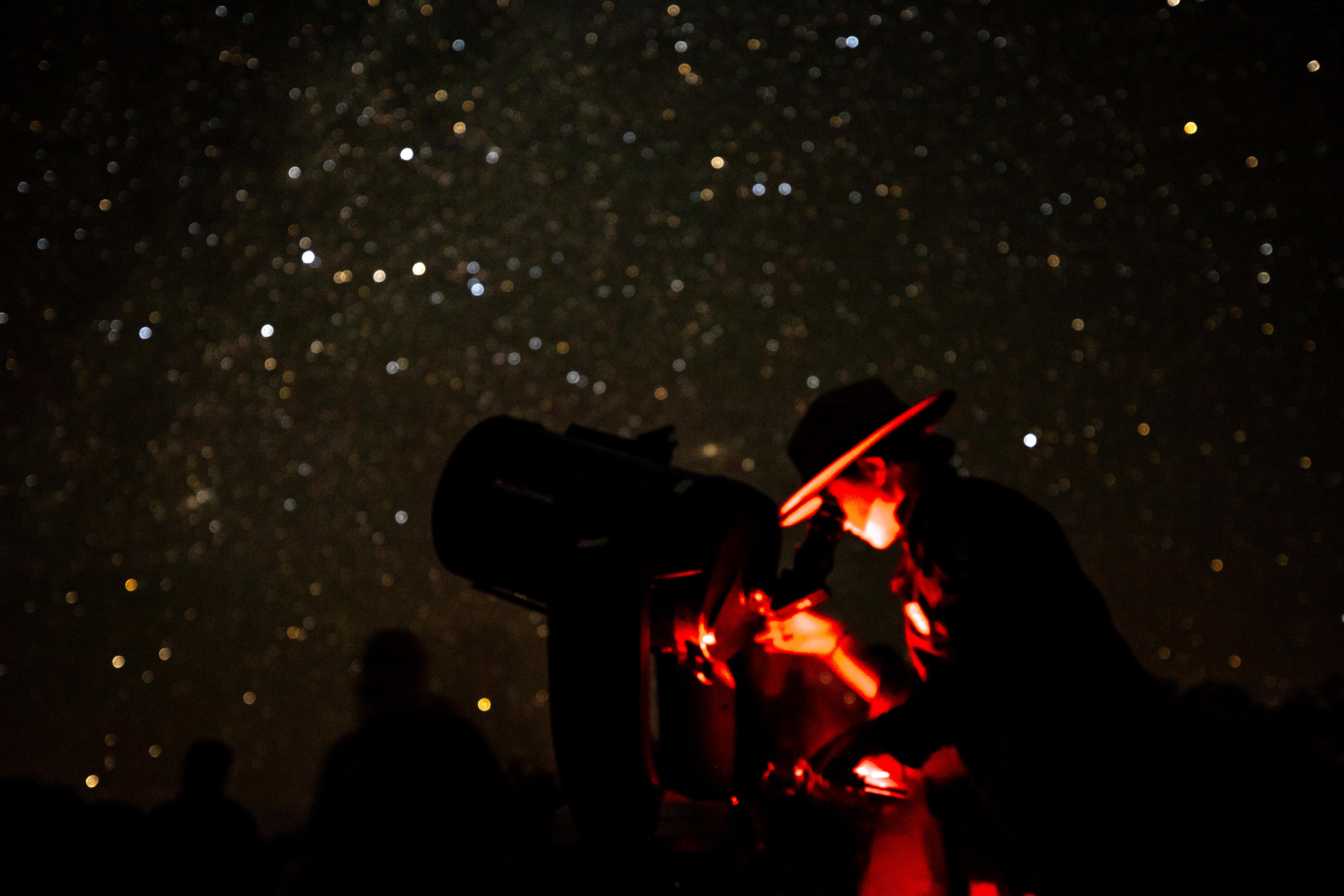A ranger, highlighted by red-light, looks into a telescope with a black backdrop filled with stars.