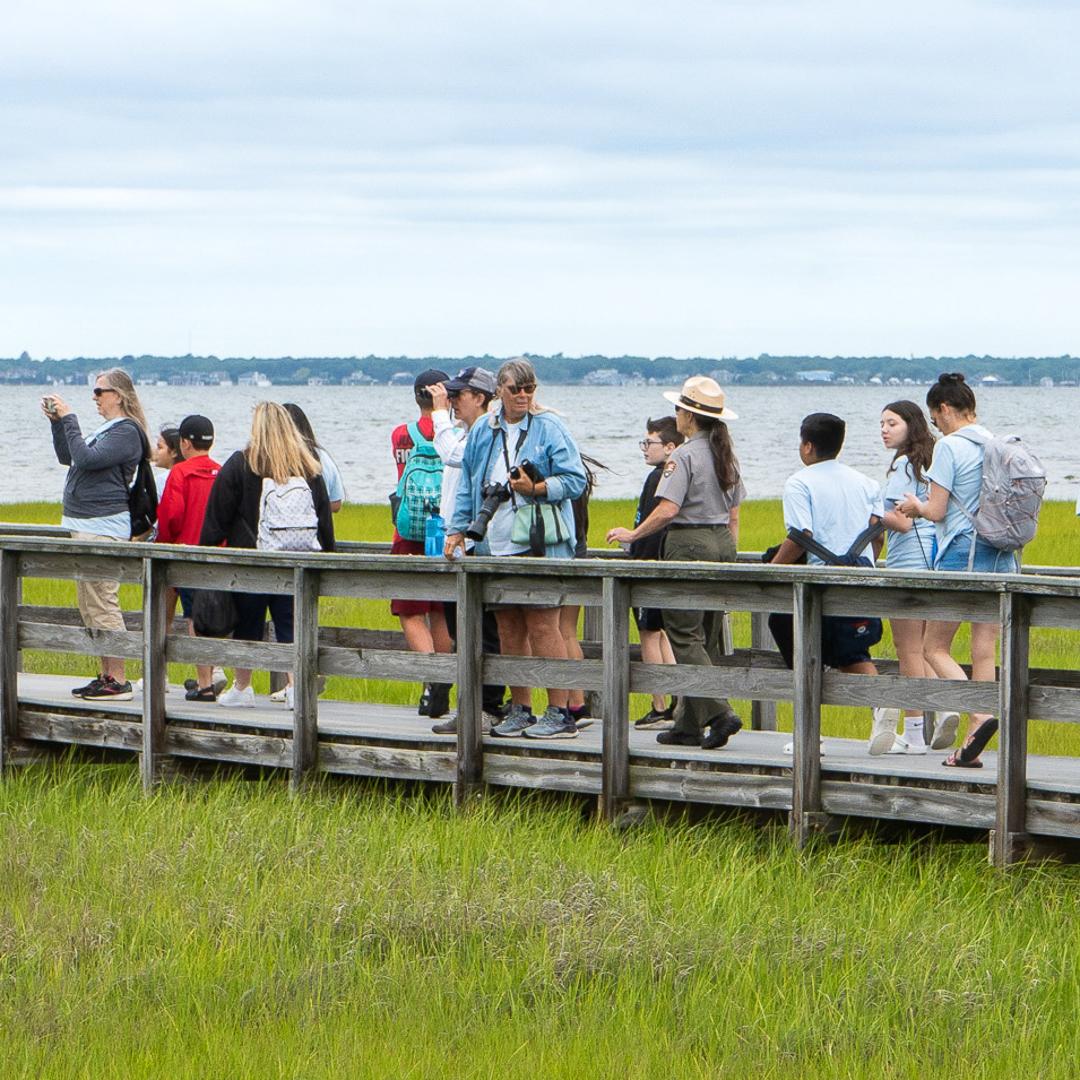 A group walks on a boardwalk over a salt marsh.