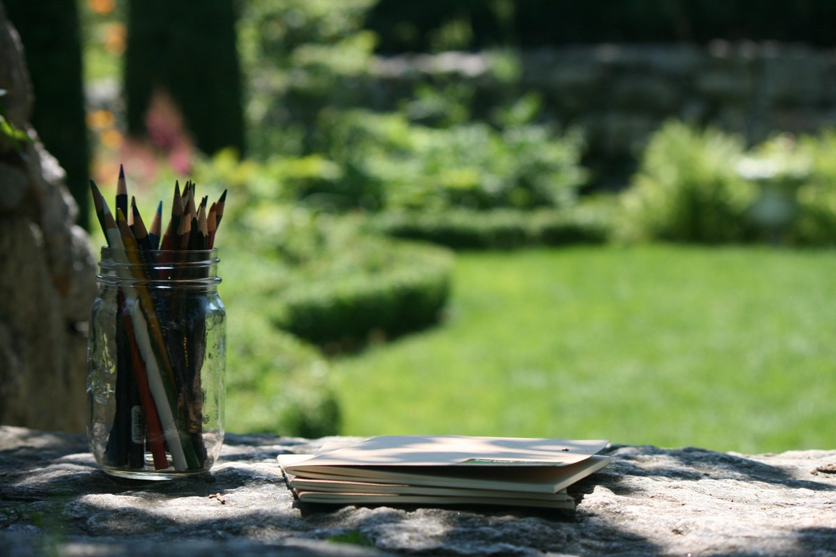 Colored pencils and journals rest on a stone wall with a garden in the background.