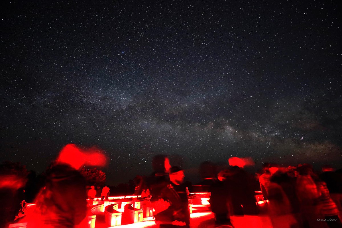 The milky way illuminates a dark sky, with visitors lined up in red light in front.