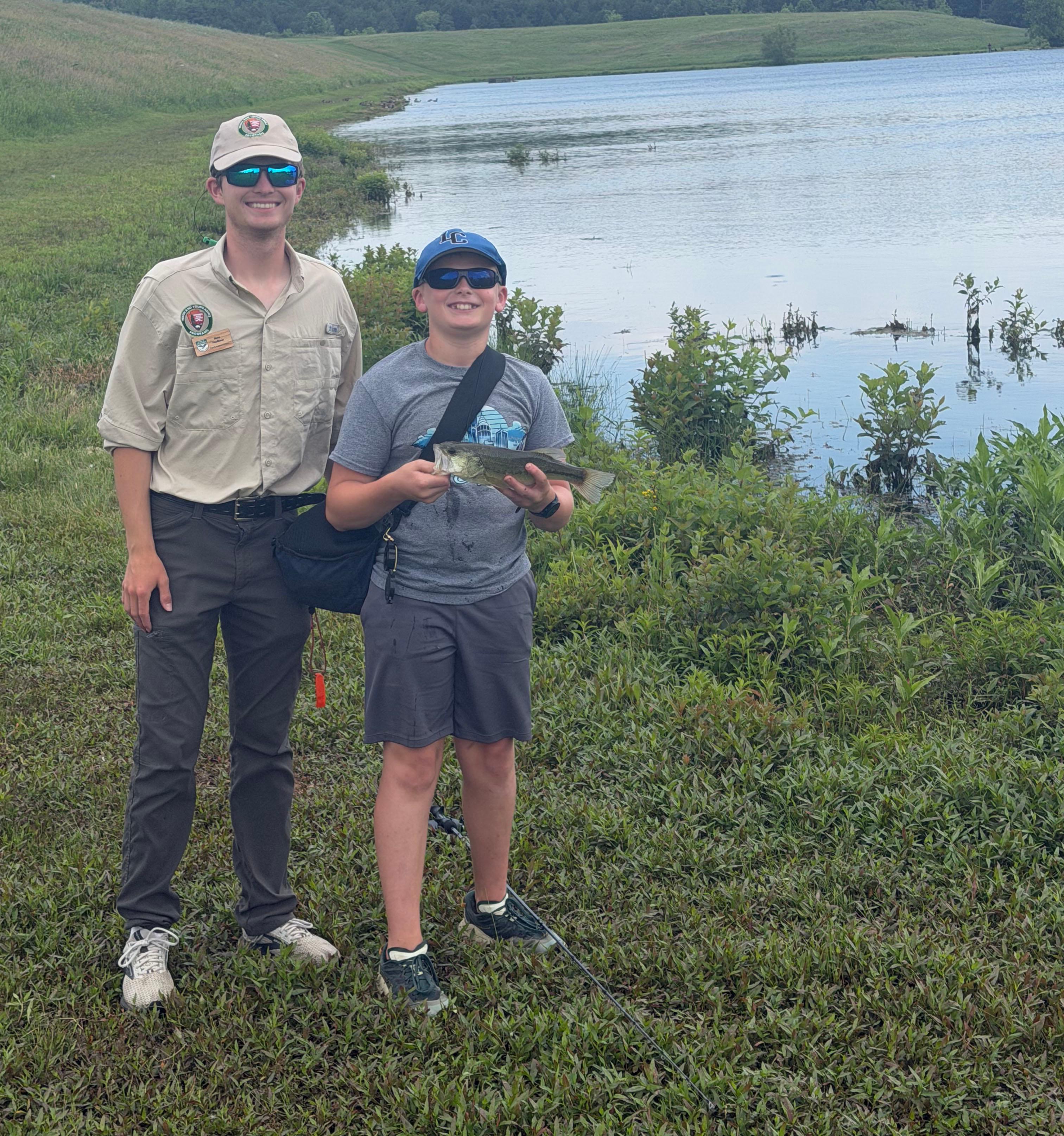 A color image showing two men, one holding a fish by a lake
