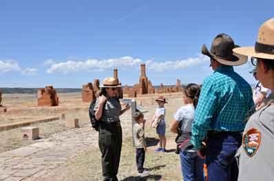 A female park ranger speaks to a group of visitors while standing in front of adobe ruins.