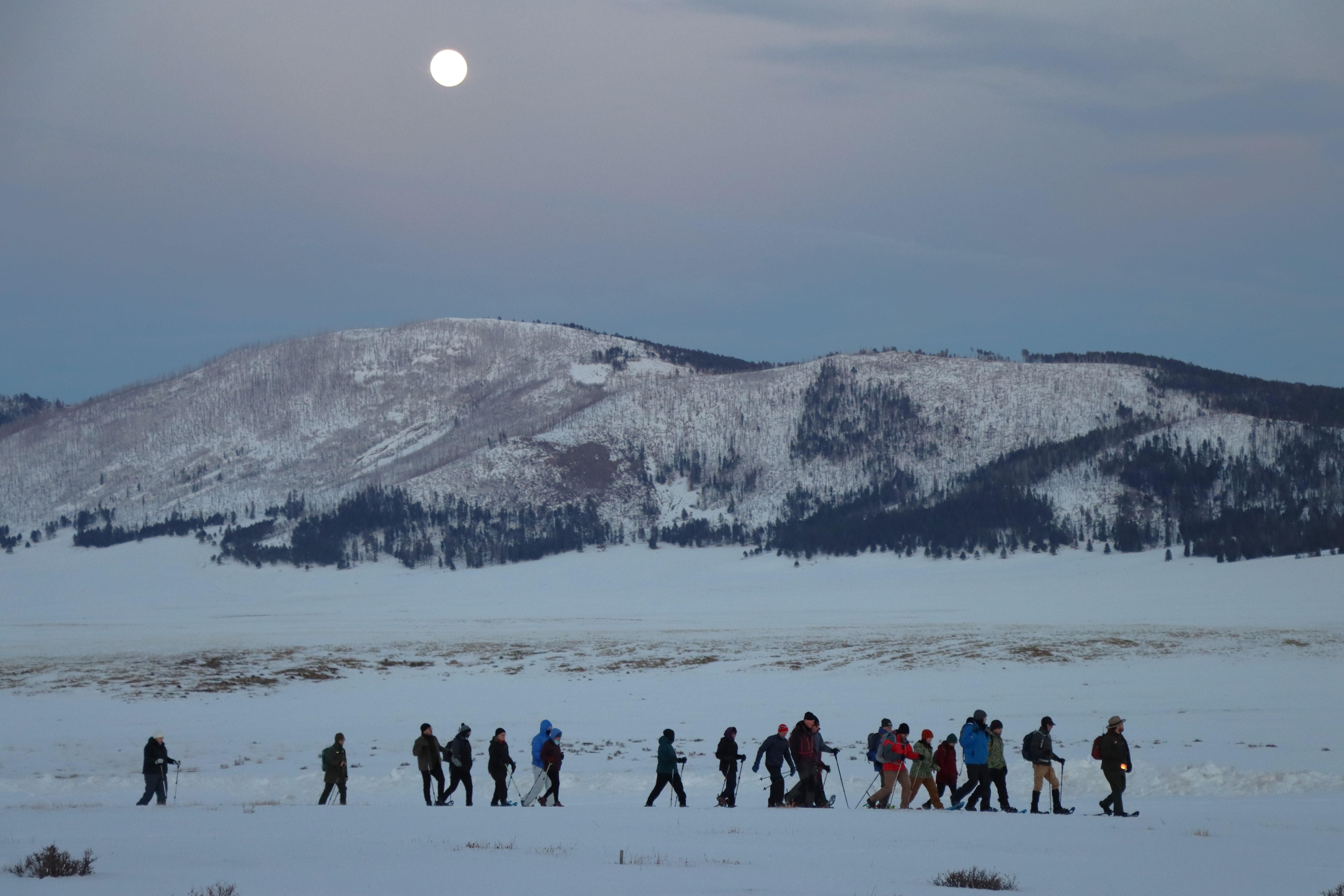 A line of snowshoers hiking through a snowy mountain landscape as the full moon rises.
