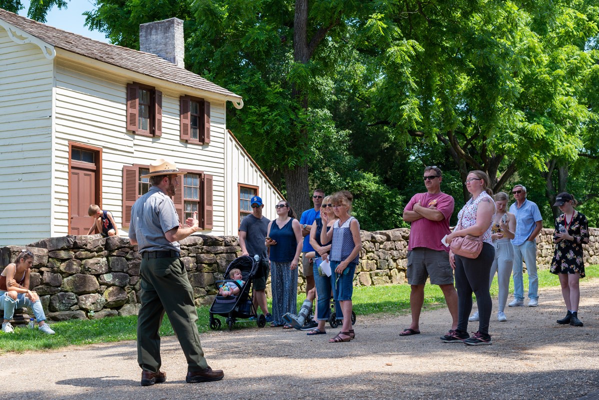 A park ranger talks to a group of visitors outside on a gravel path next to a small white house.
