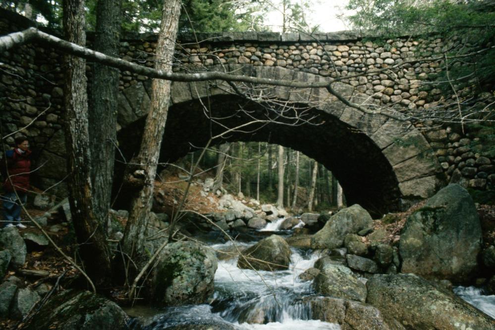 A bridge made of cobblestone spans a rocky creek surrounded by trees.