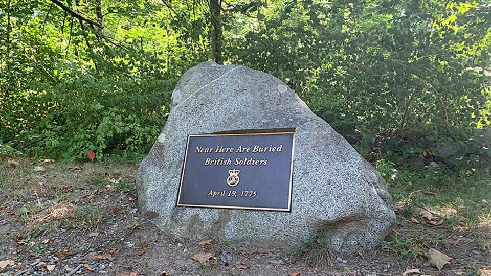 A stone sits along a dirt train. A metal sign attached to the stone marks the location of graves