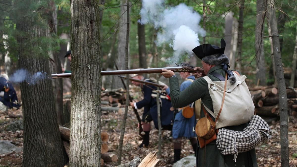 A colonial militia man fires his musket from behind a tree