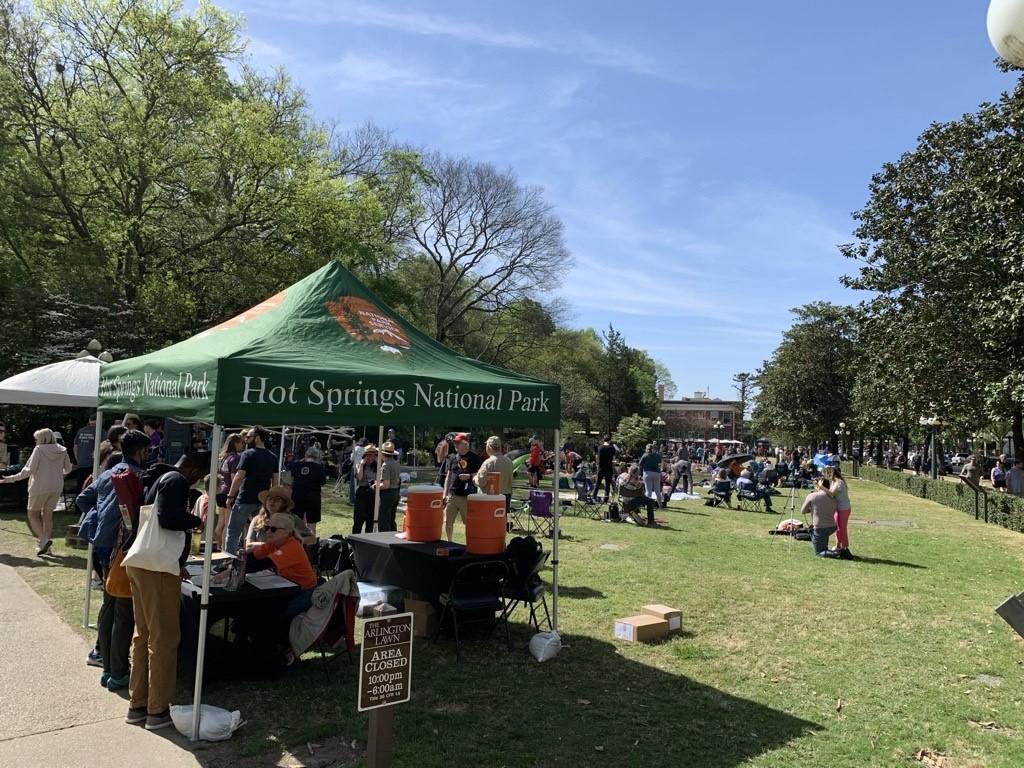 People stand under a shade tent while people in the background sit on a grassy lawn.