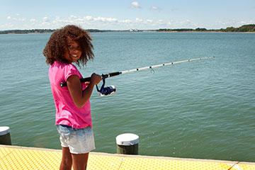 a child stands on a pier with a fishing rod, looking back at the camera.