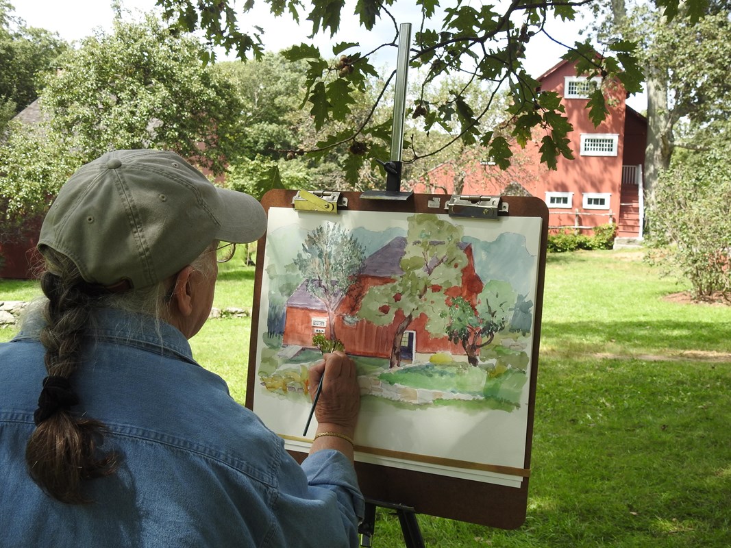 A close up image of an artist painting a red building in the background.