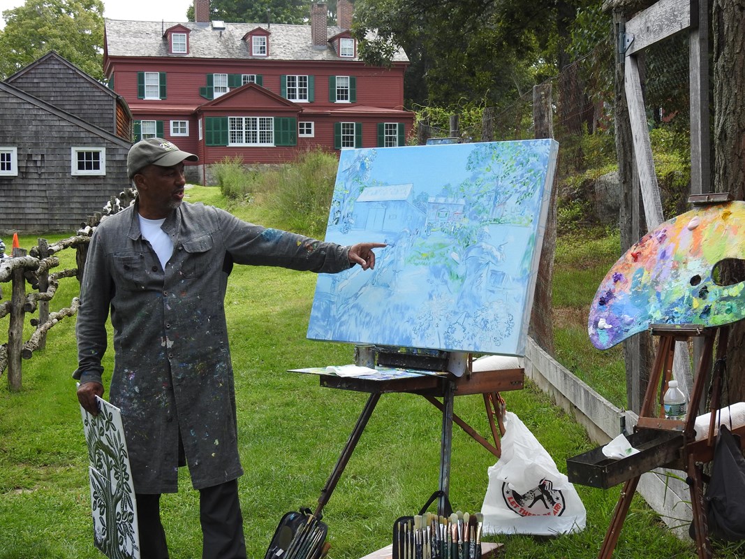 An artist stands next to his easel pointing at a piece on his canvas.