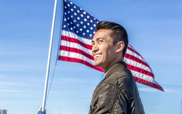 Man smiles against a background of an American flag flying in a blue sky.