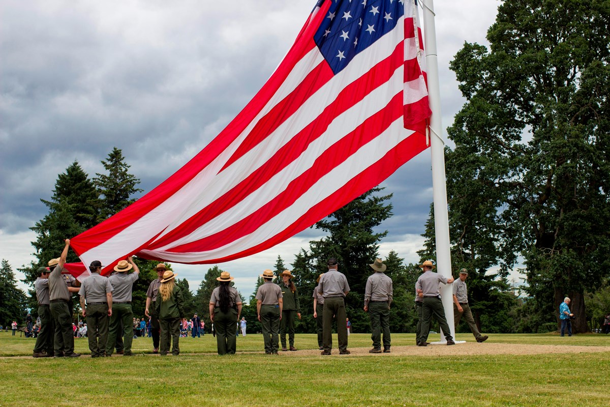 National park rangers stand on the ground as a large garrison flag is raised on a flagstaff.