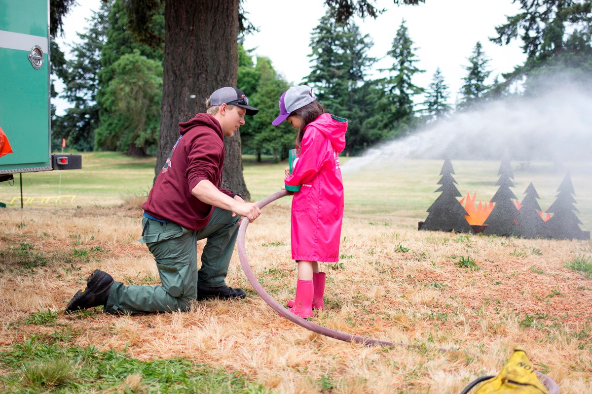 A firefighter kneels and shows a girl how to aim a hose at a pretend fire.