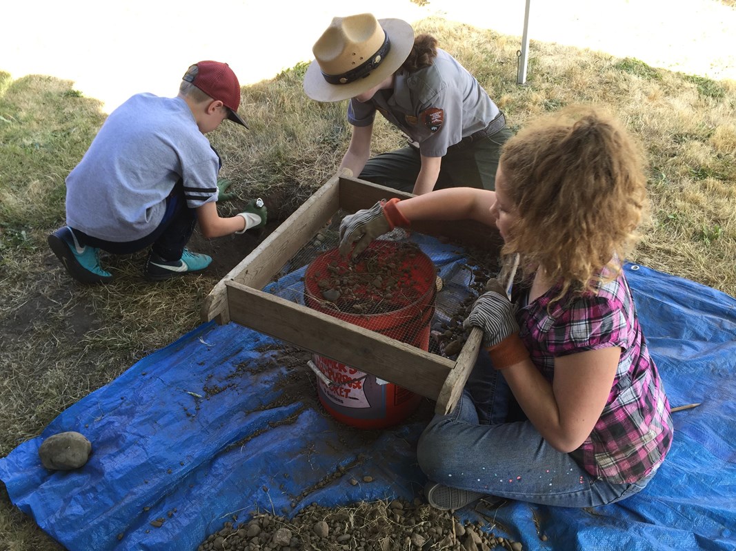 A park ranger helps one kid dig a hole in the ground while another screens dirt.