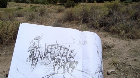 image of someone drawing sketch of a person and a wooden cart, with trail in the background