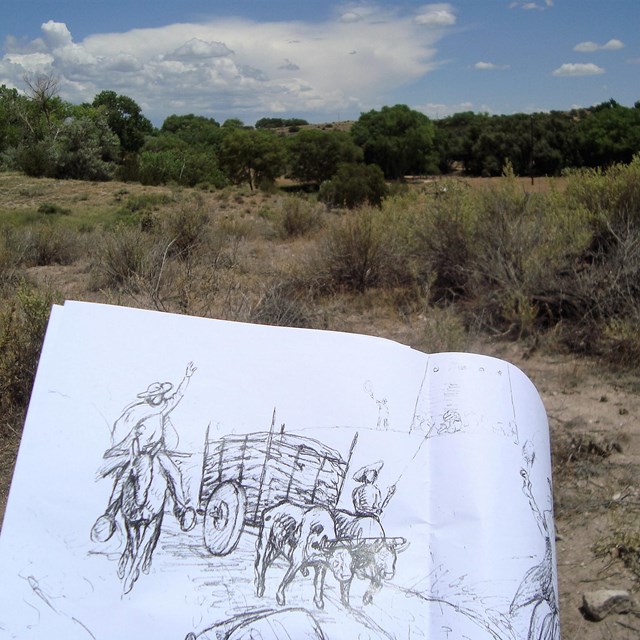 image of someone drawing sketch of a person and a wooden cart, with trail in the background